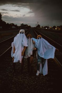 three people in white covering their faces while standing on train tracks at night with dark clouds overhead