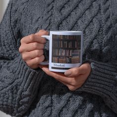 a person holding a coffee mug in their hands with bookshelves on the wall behind them