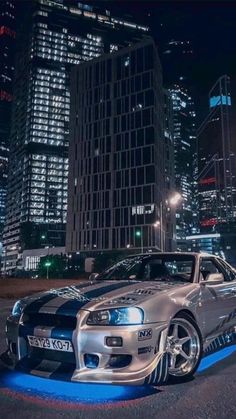 a silver sports car parked in the middle of a city at night with skyscrapers behind it