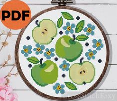 a cross stitch pattern with green apples and blue flowers on the hoop, next to a pink flower