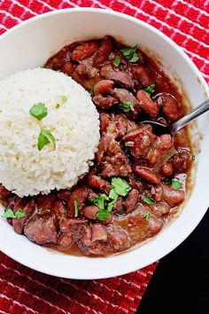 a white bowl filled with meat and rice on top of a red table cloth next to a spoon