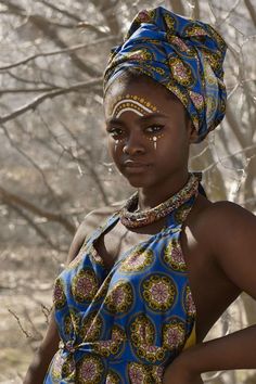 a woman in a blue and yellow dress standing next to some trees with her nose ring on