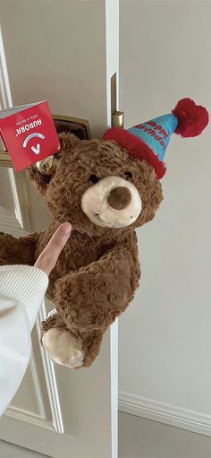 a brown teddy bear with a birthday hat hanging on a door handle and pointing at it