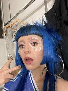 a woman with bright blue hair and makeup is holding her finger up to her face