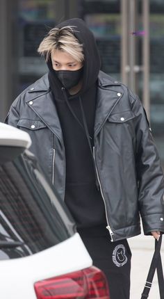 a man in a black hoodie and mask walks past a white car with his hand on the door handle