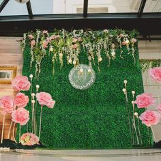 a green wall with pink flowers and greenery on the top is surrounded by white roses