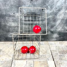 two red apples sitting in a metal basket on the floor next to a gray wall