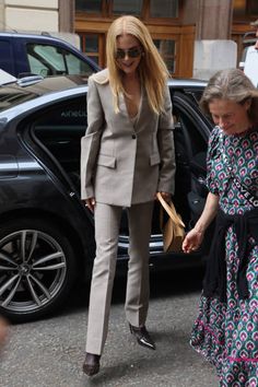 two women walking down the street in front of some cars and one is wearing a suit