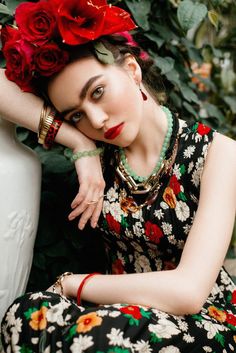 a woman with red roses on her head sitting next to a white vase and greenery