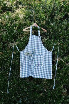 a blue and white gingham apron hanging on a wooden hanger in front of some bushes