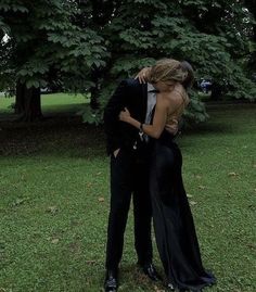 a man and woman in formal wear embracing each other on the grass with trees behind them