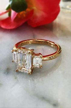 three stone diamond ring sitting on top of a marble table next to a red flower