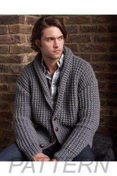 a man sitting in front of a brick wall wearing a cardigan sweater and jeans