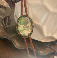 a necklace with a painting on it sitting on top of a glass plate next to a plant