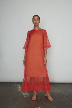 a woman standing in front of a white wall wearing an orange dress with sheer sleeves