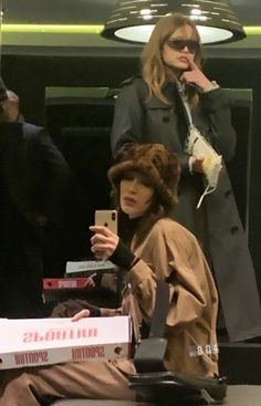 two women sitting down while one takes a photo with her cell phone and the other is wearing a fur hat