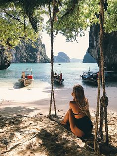 a woman sitting on the beach looking out at boats in the water and mountains behind her