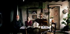 two people sitting at a table in a room with plants and pictures on the wall