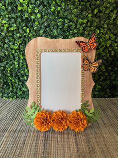 an orange flower and butterfly frame on a table next to a green bush with leaves