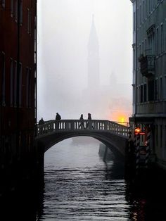 people are standing on a bridge in the foggy water near buildings and a clock tower