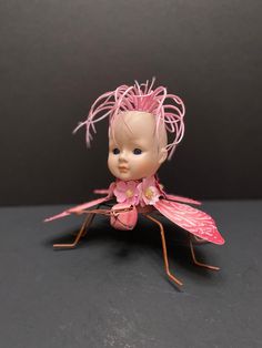 a doll with pink hair and wings sitting on a black surface in front of a gray background