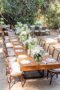 a long wooden table with white flowers and greenery on it is set for an outdoor dinner