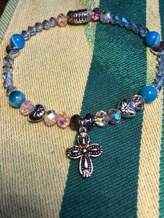 This silver-pated cross charm bracelet is filled with little surprises, like silver metal heart beads, light pink aurora borealis faceted glass beads, striped blue agate, and light blue glass beads. This bracelet is sized for a child. Silver Crystal Bracelet With Heart And Round Beads, Silver Crystal Bracelet With Heart Shaped Beads, Adjustable Silver Beaded Bracelets With Cross Shape, Adjustable Silver Cross Beaded Bracelets, Adjustable Silver Beaded Cross Bracelets, Spiritual Silver Cross Charm Bracelet, Spiritual Silver Charm Bracelet With Cross, Silver Spiritual Bracelet With Heart Beads, Blue Cross Bracelets As Gift