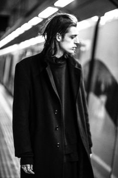 a man with long hair standing in front of a subway train at the station, wearing a black coat and scarf