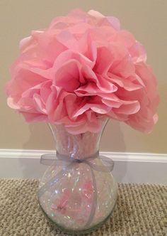 a pink flower in a clear vase on a carpeted floor with a wall behind it