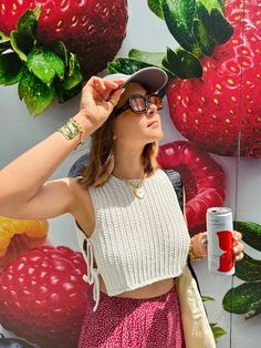 a woman wearing glasses and a hat is holding a cup in front of a wall with strawberries on it