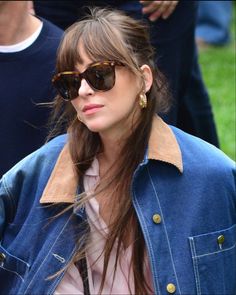 a woman wearing sunglasses and a denim jacket is looking off to the side with people in the background