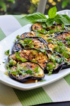 a white plate topped with eggplant covered in sauce and greens on top of a green checkered table cloth