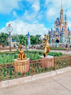 two golden statues sitting on top of a metal fence in front of a disney castle