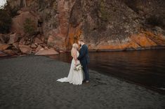 a bride and groom standing on the beach in front of some rocks at sunset time