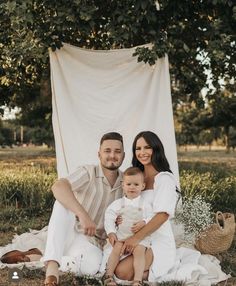 a man and woman sitting on a blanket with a baby in front of them, under a tree