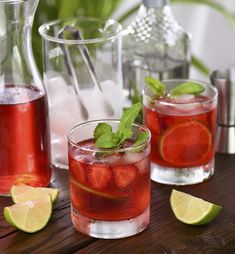 two glasses filled with red liquid and limes next to pitchers of water, lemonade and ice