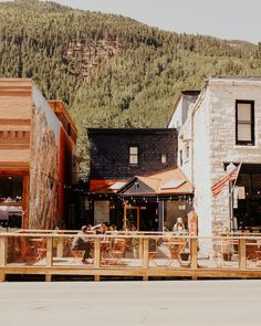 people are sitting at tables outside in front of some buildings and trees on the hill