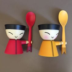 two little figurines are holding spoons on a gray surface, one is yellow and the other is red