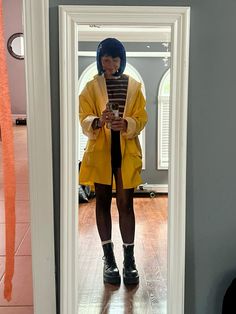 a woman standing in front of a mirror wearing a yellow coat and black boots, looking at her cell phone