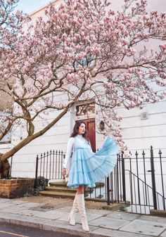 a woman in a blue dress and white boots standing on the sidewalk next to a tree