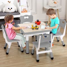 two children sitting at a table with toy cars on the table and one child writing