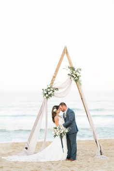 a bride and groom kissing on the beach in front of an arch decorated with flowers