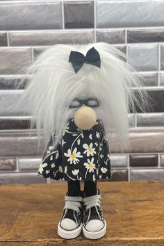 a small doll with white hair and black dress on top of a wooden table in front of a brick wall