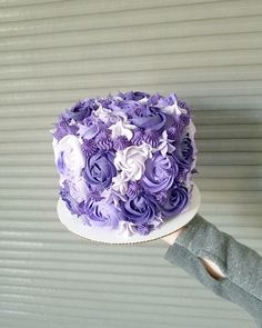 a hand holding a cake with purple and white flowers on the top, in front of a corrugated wall