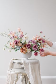 a woman arranging flowers in a vase on top of a table next to a stool