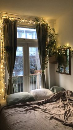 an unmade bed in front of a window with fairy lights