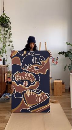 a woman standing behind a large painting in a room with wood flooring and potted plants