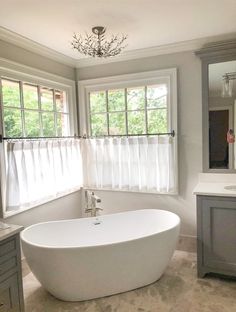 a large white bath tub sitting in a bathroom next to a sink and window with sheer curtains