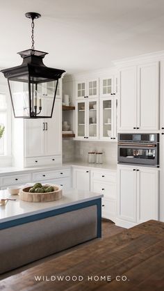 This serene white kitchen is a peaceful retreat from the hustle and bustle of everyday life. The crisp white kitchen cabinets, paired with warm wood floors, floating shelves and black hardware, creates a calming and inviting atmosphere. The large windows flood the space with natural light, making it the perfect place to be whether you're cooking, entertaining, or relaxing. Get inspired by Wildwood Home Co.'s design vision and home decor store at wildwoodhomecompany.com.