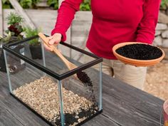 a woman holding a wooden spoon in a glass container filled with dirt and gravel on top of a table